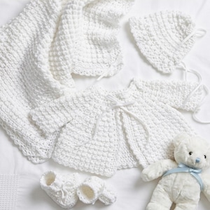 Puede incluir: Un conjunto de bebé blanco tejido a mano que incluye una manta, un gorro, una chaqueta y patucos. También se incluye un osito de peluche blanco con una cinta azul. El conjunto está hecho de hilo suave y texturizado.
