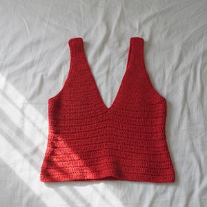 Puede incluir: Un top rojo de ganchillo hecho a mano con un escote en V profundo y tirantes anchos. El top está hecho de un hilo texturizado y se muestra sobre una superficie blanca. La prenda es sin mangas.