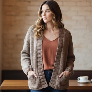 May include: A woman wearing a brown, open-front cardigan with pockets. The cardigan is made of a chunky knit material. She is wearing a rust-colored top and blue jeans. A cup of coffee and books are on a wooden table.