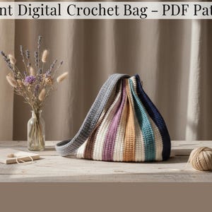 Könnte beinhalten: Gehäkelte Tasche mit Streifen in verschiedenen Farben, darunter Blau, Lila und Beige. Die Tasche hat einen grauen Riemen und wird auf einer Holzoberfläche präsentiert. Das Bild zeigt auch eine Vase mit Trockenblumen und ein Knäuel Garn.