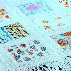 Puede incluir: Primer plano de un edredón hecho a mano con un diseño de patchwork. El edredón presenta cuadrados de tela con estampados florales en varios colores, incluyendo azul, naranja y rosa, sobre un fondo blanco de ganchillo.