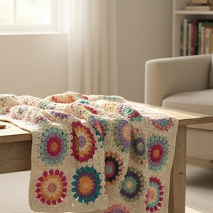 May include: A cream-coloured, crocheted blanket with colourful floral patterns is draped over a wooden coffee table. The blanket features vibrant hues of pink, orange, blue, and purple. A cup of tea sits on the table.