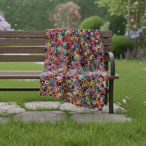 May include: A wooden park bench with a colorful floral blanket draped over it. The blanket features a pattern of various colored flowers. The bench is set on a stone path in a grassy area, with trees and flowers in the background.