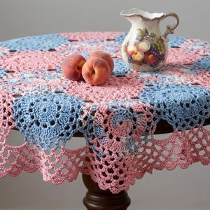 May include: A round, crocheted tablecloth in a patchwork design of pink and light blue squares. A decorative ceramic pitcher with fruit illustrations and three peaches sit on the table. The tablecloth has a scalloped edge.