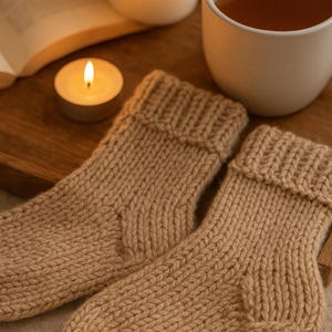 Op de afbeelding: Een paar beige gebreide sokken met geribde manchetten, op een houten dienblad. Een brandende kaars, een kopje thee en een open boek staan ook op de dienblad, wat een gezellige, ontspannende scène creëert.