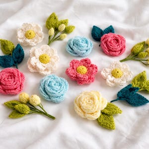 May include: A collection of handmade crochet flowers in various colors, including pink, blue, white, and yellow. The flowers are arranged with green leaves and buds on a white background, showcasing a craft project.