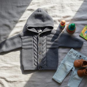 Peut inclure: Un cardigan gris à capuche pour bébé avec un motif en tricot torsadé, une fermeture à boutons, un jean bleu clair assorti et des chaussures en cuir marron. Un jouet à empiler coloré et un petit livre sont également présents.
