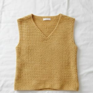 Op de afbeelding: Mosterdgele gebreide vest met een V-hals. De mouwloze top is gemaakt van een getextureerde gebreide stof. Het vest is effen van kleur en heeft een klein label bij de halslijn. Het kledingstuk wordt gepresenteerd op een witte ondergrond.