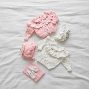Op de afbeelding: Een collectie babykleding in zachtroze en wit, waaronder een vest, muts en babyschoentjes. De kledingstukken hebben delicate breipatronen en ruches. De set is gerangschikt op een witte ondergrond.