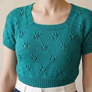 May include: Teal-colored short-sleeved sweater with a square neckline. The knit top features a floral pattern throughout the bodice and sleeves. The bottom hem and neckline are ribbed. The sweater is a cropped style.
