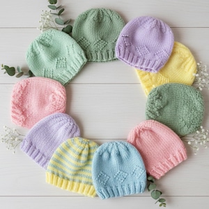 May include: A collection of colorful, knitted beanies in pastel shades of green, pink, purple, yellow, and blue. The hats are arranged in a circle, with sprigs of greenery and white flowers interspersed between them. The hats have various knit patterns.