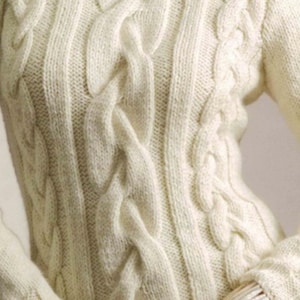 May include: Cream-colored cable knit sweater. The close-up shows the intricate woven pattern of the sweater, with thick, raised cables running vertically and diagonally across the front and sleeves. The sleeves are long and the neckline is a crew neck.