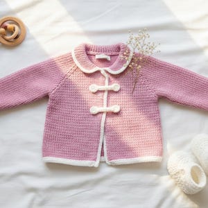 Może przedstawiać: Jasnoróżowy sweterek dla dziecka z białymi wykończeniami i zapięciami na kołki. Sweterek ma biały kołnierz i mankiety. Drewniana grzechotka i para białych bucików dla dziecka również znajdują się na zdjęciu.