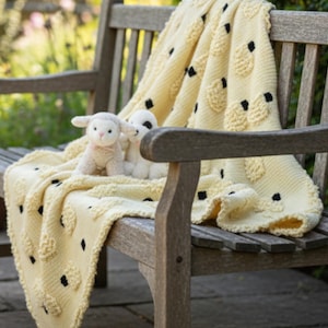 Könnte beinhalten: Eine cremefarbene Strickdecke mit Schafmuster, drapiert über einer Holzbank. Zwei Plüschlamm-Spielzeuge liegen auf der Decke. Die Decke hat ein strukturiertes Design mit schwarzen Akzenten und schafft eine gemütliche Szene.