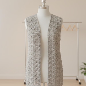 May include: A light gray, open-front crochet vest displayed on a mannequin. The vest features a detailed, textured stitch pattern and a long, sleeveless design. A clothing rack and a potted plant are visible in the background.