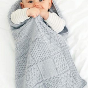Könnte beinhalten: Ein Baby, eingehüllt in eine hellgraue Kapuzenstrickdecke. Die Decke hat ein strukturiertes, geometrisches Muster. Das Baby trägt ein weiß-grau gestreiftes Langarmshirt.
