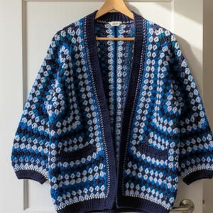 May include: A blue, white, and navy blue crocheted cardigan with a geometric pattern. The open-front sweater has two front pockets and is hanging on a wooden hanger against a white door.