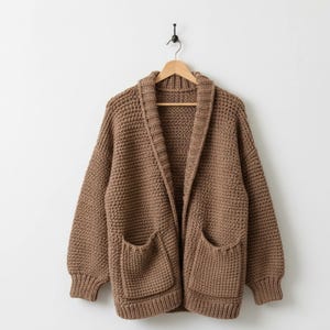Può includere: Un cardigan marrone a maglia grossa, con collo a scialle e due tasche frontali, è appeso a una gruccia di legno. Il maglione ha una vestibilità ampia e polsini a coste. Lo sfondo è un muro bianco.