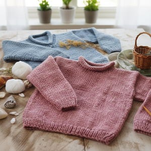 Puede incluir: Primer plano de dos jerséis de punto, uno rosa empolvado y otro azul claro, con un ovillo de lana y agujas de tejer. Los jerséis están sobre una superficie de madera, con elementos decorativos como conchas y una pequeña cesta.