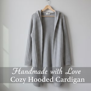 Peut inclure: Un cardigan à capuche gris clair suspendu à un cintre en bois. Le pull à devant ouvert a une texture en tricot côtelé et des manches longues. Le texte "Handmade with Love Cozy Hooded Cardigan" est affiché en bas de l'image.