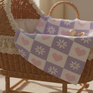 May include: A wicker bassinet with a floral patterned blanket. The blanket features a checkerboard design with lavender squares, white squares, pink hearts, and white flowers. The bassinet has a lace trim.