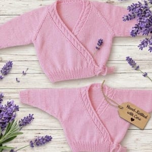 May include: Two pale pink, hand-knitted baby cardigans with a wrap-style design. The cardigans have long sleeves and a braided detail along the neckline. A wooden tag reads "Hand-Knitted with Care". Lavender sprigs are arranged around the cardigans.