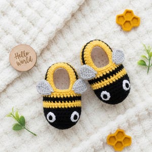 Puede incluir: Un par de patucos de bebé hechos a mano de ganchillo con diseño de abeja. Los patucos son amarillos y negros con alas grises y ojos blancos. En la imagen también hay un disco de madera con las palabras "Hello World".