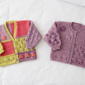 May include: Two colorful, hand-knitted cardigans. One is a patchwork design in pink, yellow, and lavender, adorned with flower appliqués. The other is a solid lavender with floral embellishments and button closures. Both are on a white background.
