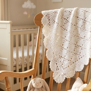 Könnte beinhalten: Eine cremefarbene, gehäkelte Babydecke mit gewelltem Rand, die über der Rückenlehne eines Holzschaukelstuhls drapiert ist. Die Decke hat ein geometrisches Lochmuster. Ein Stoffhase und ein Kissen befinden sich am Fuß des Stuhls. Im Hintergrund ist ein Kinderbett zu sehen.