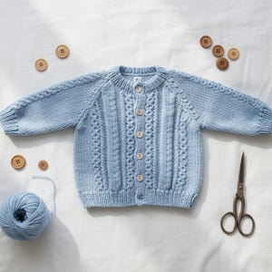 May include: A light blue, hand-knitted baby cardigan with a cable knit design. The cardigan has wooden buttons and is surrounded by wooden buttons, a ball of yarn, and scissors. The cardigan is a soft, cozy garment.
