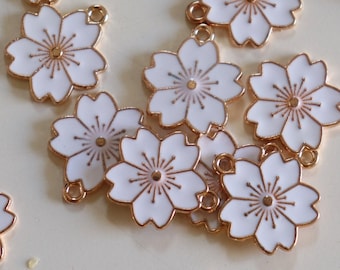 May include: A collection of gold-toned metal charms shaped like white cherry blossoms. Each charm has a small loop for attaching to jewelry or other crafts.