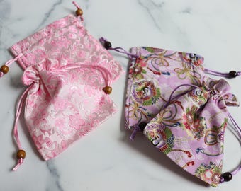 May include: Four small drawstring bags in pink and purple hues. Two bags feature a solid pink floral design, while the others display a floral and bird pattern. Each bag has a drawstring closure with wooden beads.