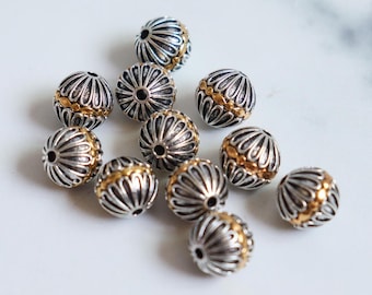 May include: A collection of silver-colored beads with gold-colored accents. The beads have a detailed, textured design and are scattered across a white surface. The beads are round and appear to be jewelry components.