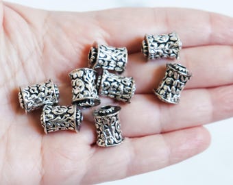 May include: A collection of silver-colored, cylindrical beads with intricate floral patterns. The beads are held in a hand, showcasing their detailed design and metallic sheen. These decorative beads are suitable for jewelry making.