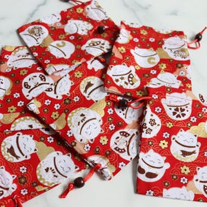 May include: Several red drawstring pouches with a repeating pattern of white and gold lucky cats, flowers, and clouds. The pouches have red drawstrings with wooden beads. The pouches are made of fabric.