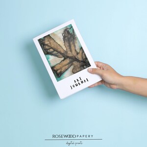 May include: A rectangular art journal with a white cover featuring a central illustration of a brown, dried leaf against a teal and cream background. The words ART JOURNAL are printed in black, sans-serif capital letters below the illustration.