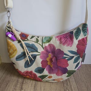 Könnte beinhalten: Eine halbmondförmige Umhängetasche mit floralem Muster und cremefarbenem Riemen. Die Tasche zeigt gestickte Blumen in Rosa, Lila und Rot, mit grünen Blättern auf cremefarbenem Hintergrund. Ein kleines lila Accessoire hängt am Riemen.