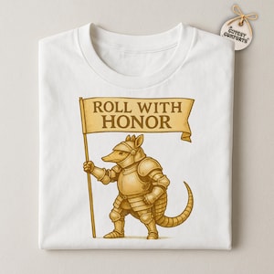 May include: White t-shirt featuring a cartoon armadillo in golden armor holding a banner that reads "ROLL WITH HONOR." The design is detailed with shading and highlights. A small tag with the text "CUTSEY COMFORTS" is attached.