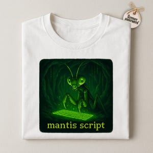 May include: White t-shirt featuring a green illustration of a praying mantis in a cave, typing on a glowing keyboard. The text "mantis script" is below the image. The shirt has a tag that says "Cutesy Comforts".