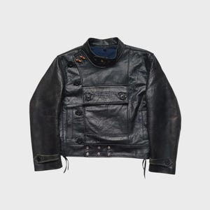Puede incluir: Chaqueta de cuero negro con cuello alto y múltiples bolsillos. La chaqueta presenta una disposición de botones única y detalles decorativos. El cuero parece desgastado, dándole un aspecto vintage. La chaqueta tiene un corte entallado y un diseño clásico.