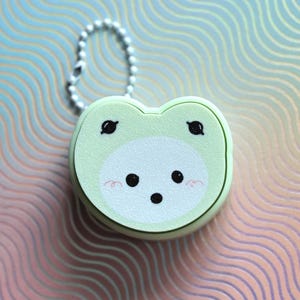 May include: A light green frog-shaped keychain with a white face, black eyes, and rosy cheeks. The keychain has a silver chain and a round, compact design. The background has a wavy pattern in shades of blue, pink, and gold.
