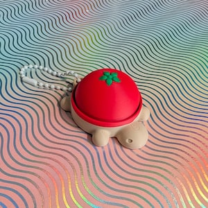 Op de afbeelding: Een novelty sleutelhanger met een schildpad ontwerp. De schildpad heeft een felrode schild, met een groen bladeren ontwerp, en het lichaam is lichtbeige. De sleutelhanger bevat een witte kralenketting.