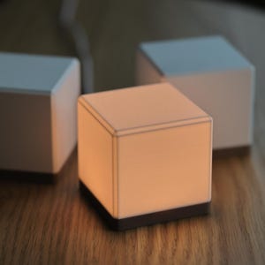 Puede incluir: Tres lámparas blancas en forma de cubo, una iluminada con un brillo cálido, sobre una superficie de madera. La lámpara encendida tiene una base oscura y un contorno definido, mientras que las otras dos están apagadas. Se ve un cable.
