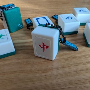 May include: White and teal Mahjong tile keychains with black chain. The tiles feature various symbols in red, blue, and green. The keychains are on a wooden surface. The tiles are cube-shaped.