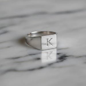 May include: A silver signet ring with a square face. The ring features the letter "K" engraved on the face. The ring is highly polished and reflects the light. The ring is set against a marble background.