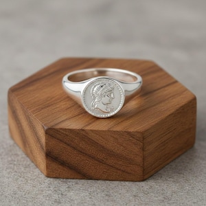 May include: A silver ring with a circular face featuring a detailed profile of a person in a laurel wreath. The ring is set on a wooden, hexagonal display stand.