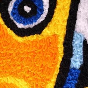 Pu&ograve; includere: Primo piano di un'opera d'arte tessile vibrante e strutturata. Il design presenta un motivo audace e astratto con una forma centrale a forma di occhio in blu, nero e bianco, circondata da giallo brillante e arancione. L'opera ha un aspetto tattile e fatto a mano.