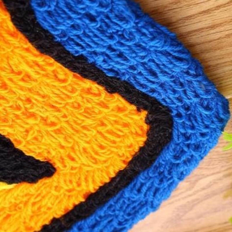 Pu&ograve; includere: Primo piano di un tappeto colorato fatto a mano con una superficie strutturata. Il tappeto presenta un design vibrante con filato giallo, nero e blu. Il tappeto &egrave; probabilmente realizzato in lana o materiale simile ed &egrave; posto su una superficie di legno.