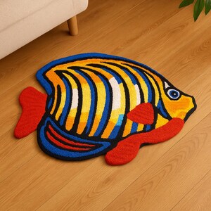 Pu&ograve; includere: Un tappeto a forma di pesce dai colori vivaci. Il tappeto presenta un contorno blu, strisce gialle e blu e pinne rosse. &Egrave; realizzato in un materiale morbido e strutturato ed &egrave; posizionato su un pavimento in legno.