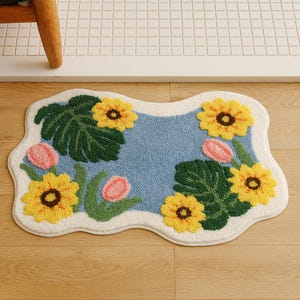 Peut inclure: Un tapis de bain décoratif à thème floral avec une bordure ondulée blanc cassé. Le tapis présente un centre bleu clair avec des tournesols jaunes, des tulipes roses et des feuilles vertes. Conçu pour un sol en bois.